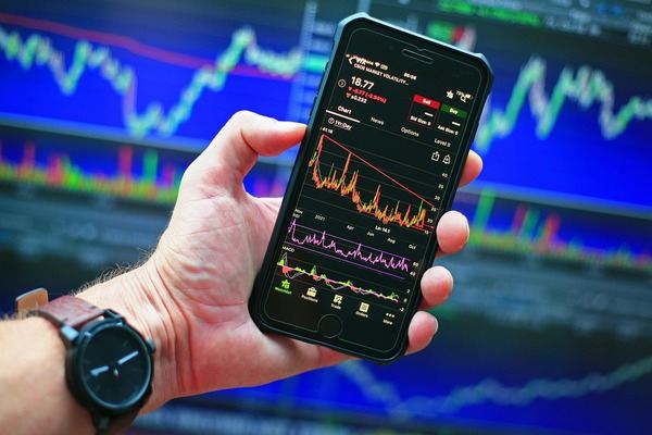 Hand holding a smartphone with a crypto price chart in front of trading screens