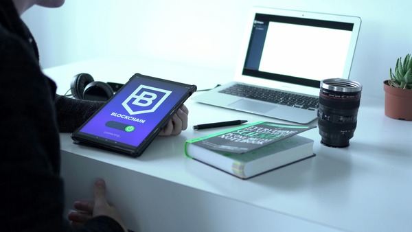 Desk with a tablet showing a blockchain app, a laptop and a book
